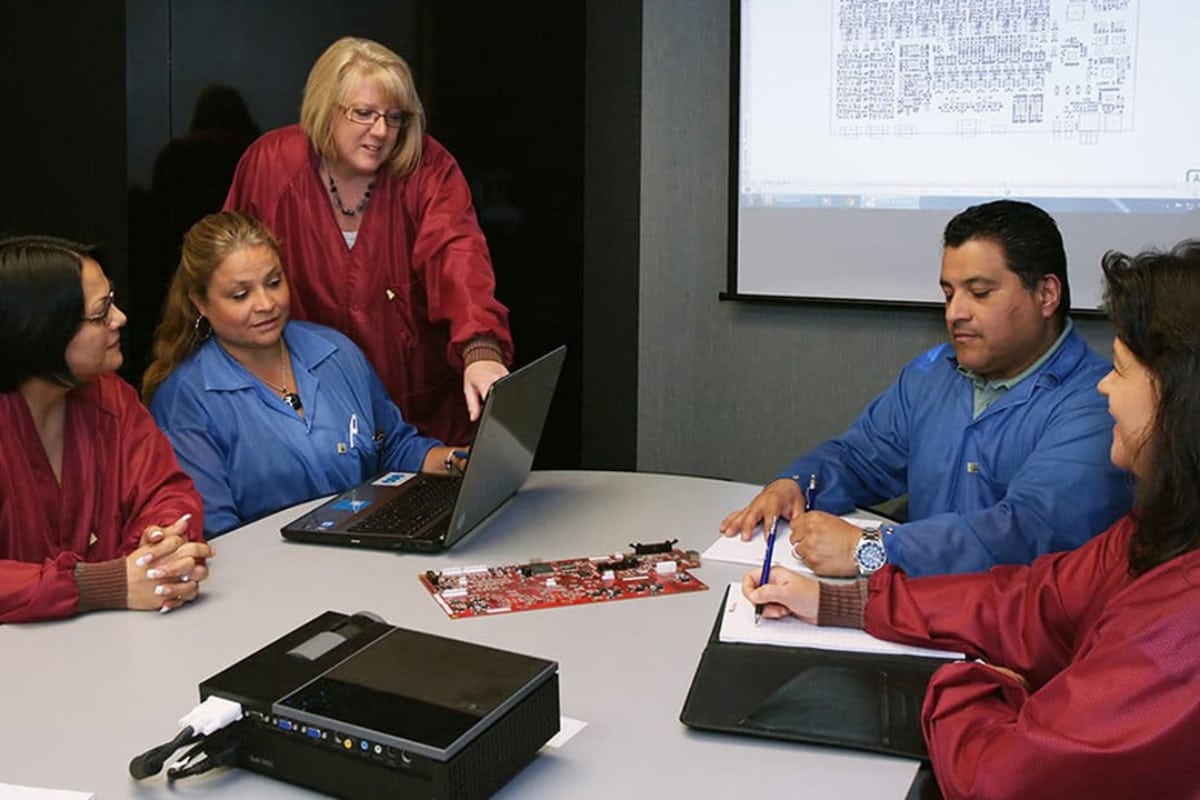 Federal Elec Blog-20 Photo-02 Five Federal Electronics team members review a circuit board and technical information around a conference table.